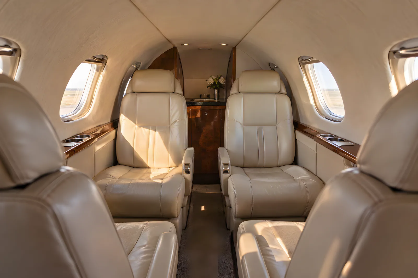 Cessna Citation I cabin interior illustration: warm natural light fills a low, narrow Cessna Citation I cabin with six cream leather armchairs, upright rounded rectangular windows and polished wood veneer, viewed aft from seated eye level down the aisle.