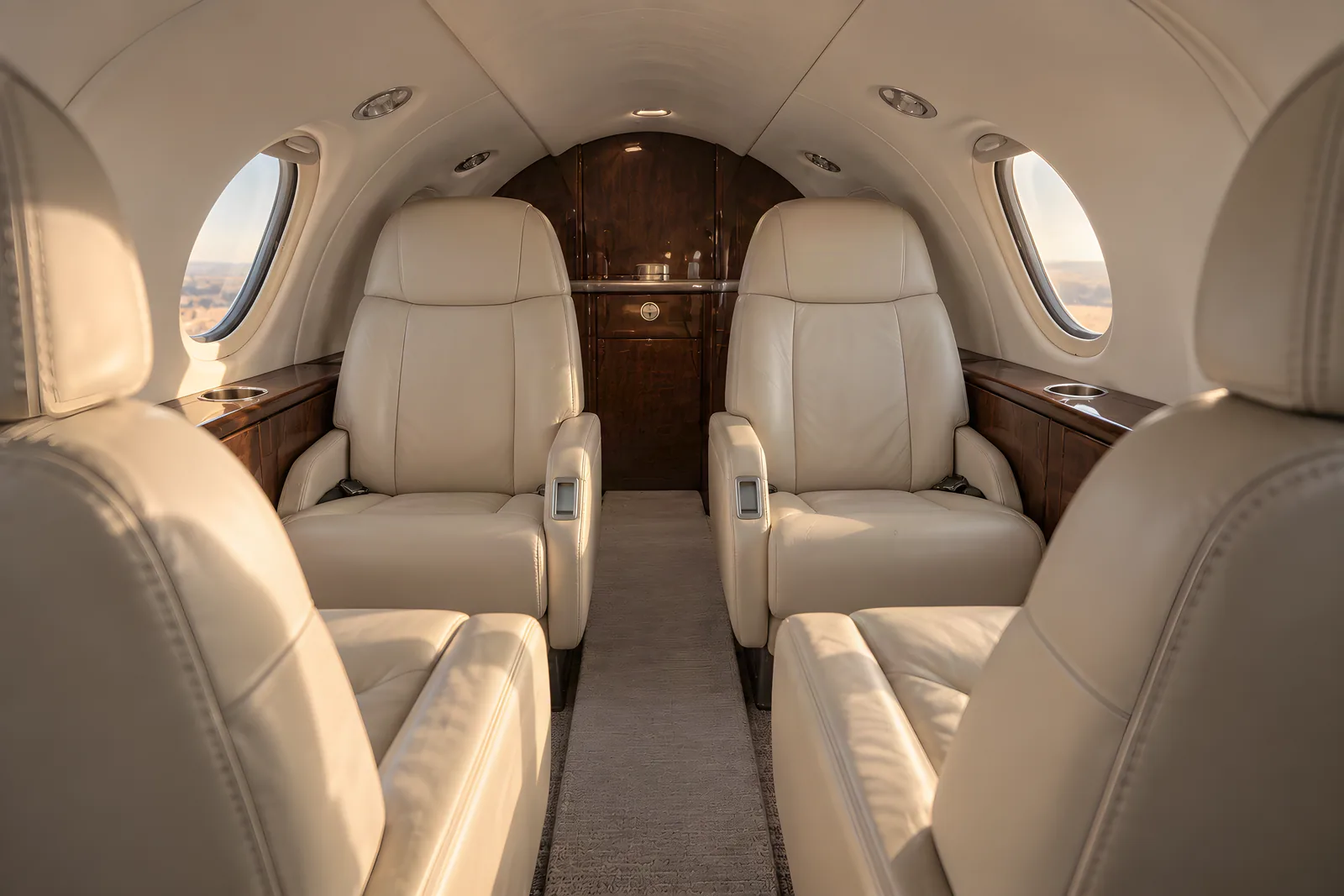 Cessna Citation Mustang cabin interior illustration: warm natural light illuminates four cream leather club seats, wood veneer and horizontal oval windows in the low, narrow Citation Mustang cabin, viewed aft along the aisle.