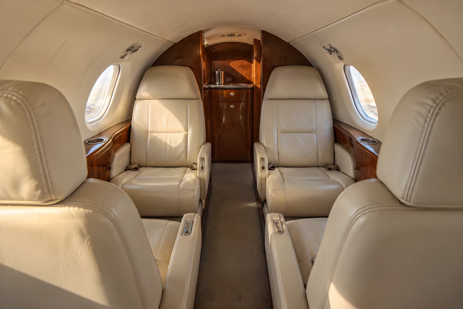 Dassault Falcon 10 cabin interior illustration: warm natural light illuminates cream leather club seats, a two-place rear bench and wood veneer in the low, narrow Falcon 10 cabin, viewed aft along the aisle through small oval windows.