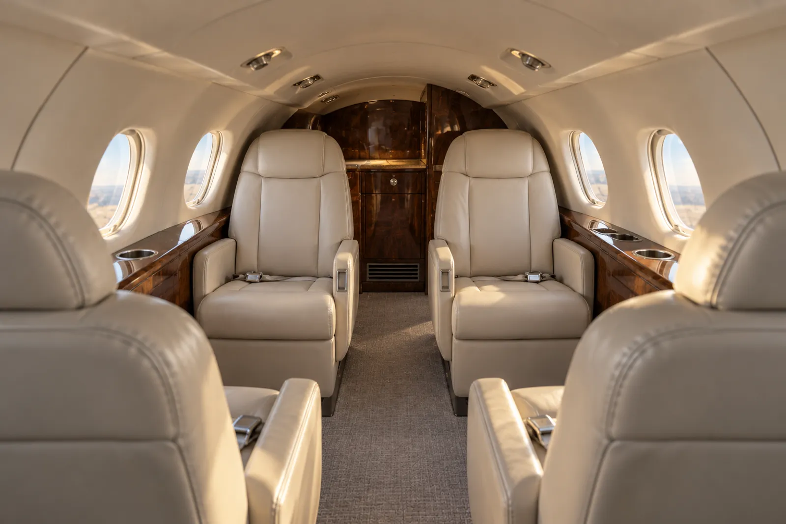 Warm natural light fills a low, narrow Beechjet 400A cabin with seven cream leather chairs, round windows, wood veneer cabinetry and a flat central aisle viewed looking aft.