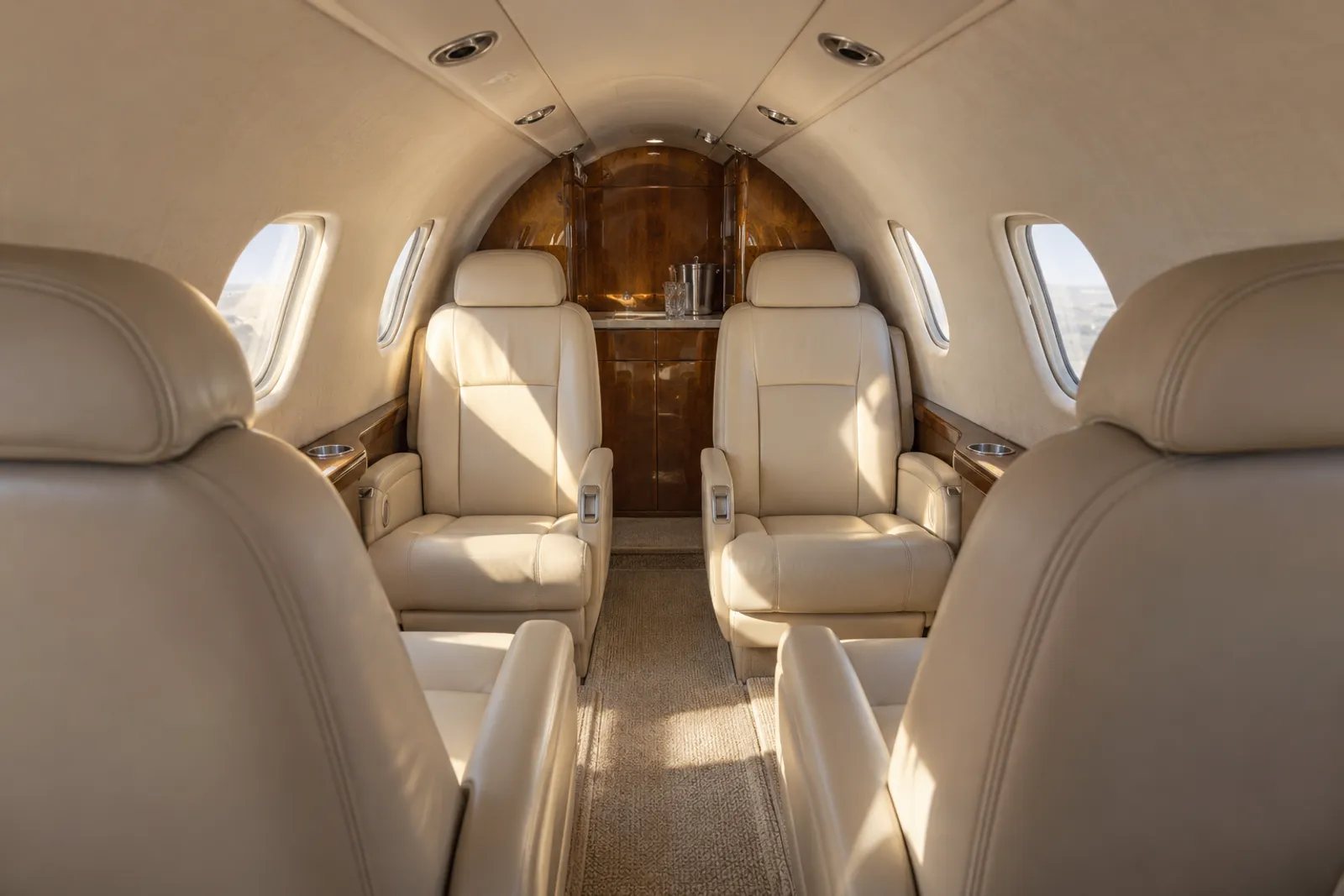 Warm sunlight fills a low, narrow Cessna Citation CJ2 cabin viewed aft along the aisle, with six cream leather armchairs, compact oval windows, wood veneer trim and an aft lavatory door.