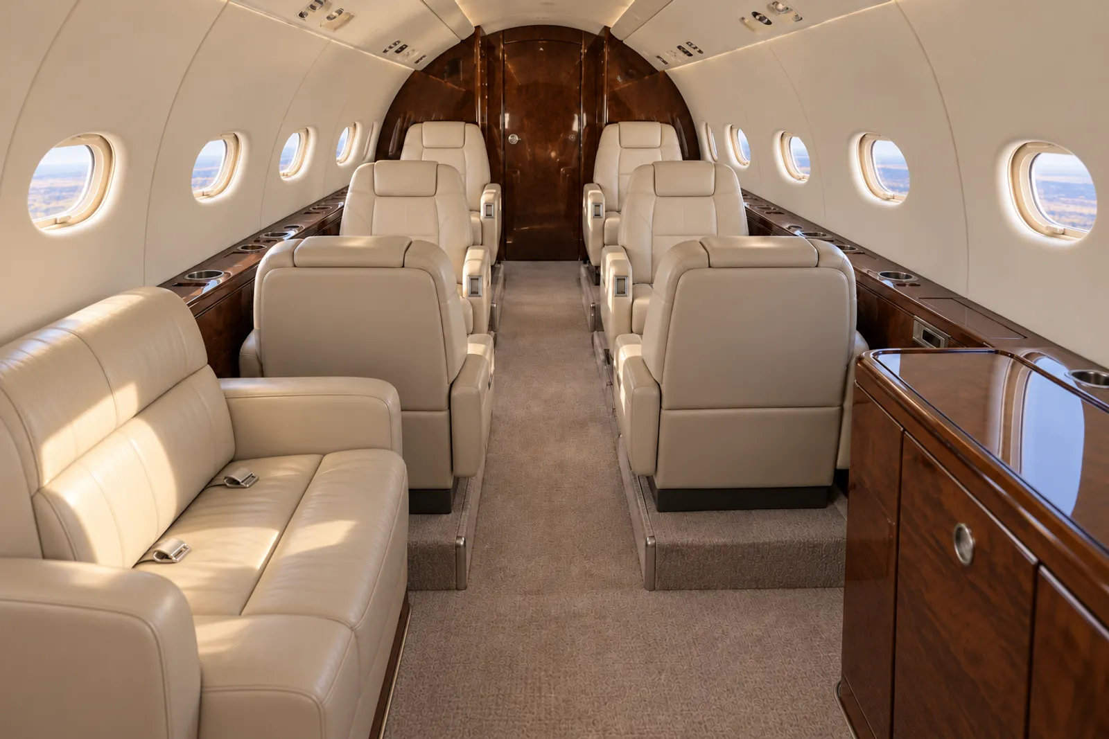 Warm natural light illuminates cream leather club seats and a two-place divan in the compact curved Citation Excel cabin, with oval windows, wood veneer and a dropped aisle leading aft to the lavatory.