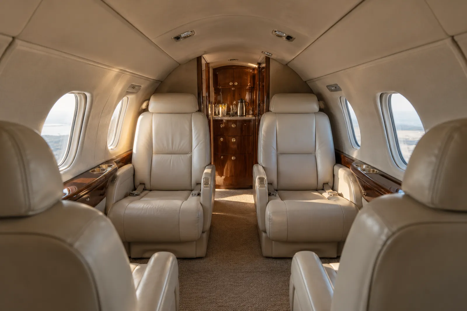 An eye-level view looking aft through the low, narrow Cessna Citation V cabin shows eight cream leather seats in two clubs, small oval windows, wood veneer cabinetry and an aft lavatory door in warm natural light.