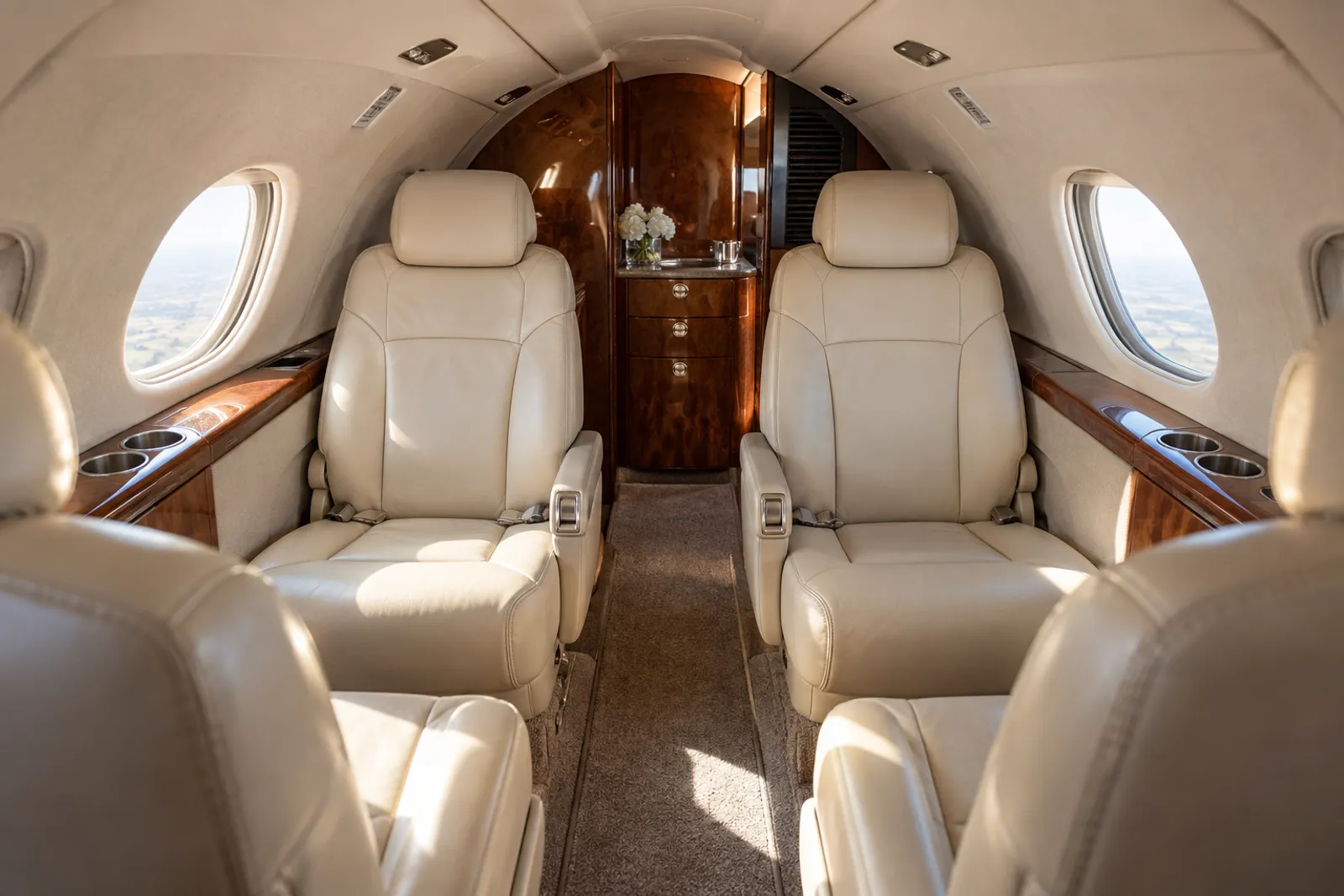 Warm natural light illuminates cream leather club chairs, wood veneer and vertical oval windows in the low, narrow CitationJet CJ1/M2 cabin, viewed aft down the aisle.