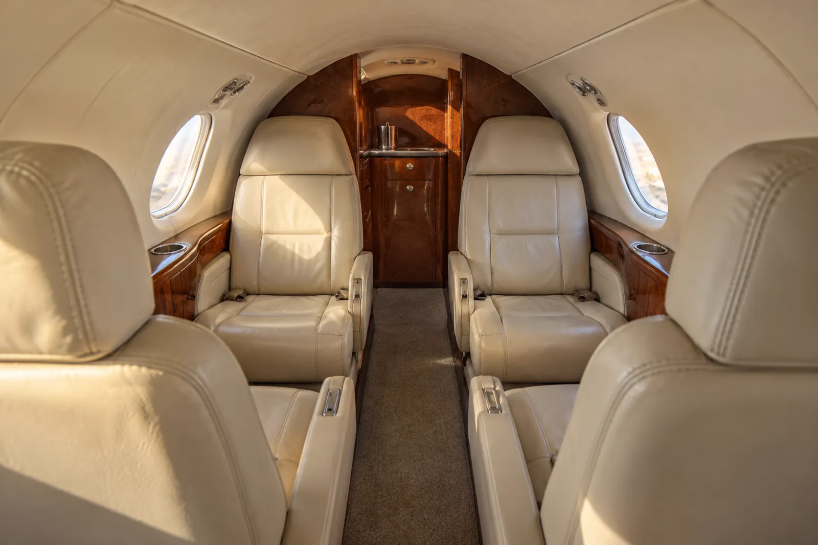 Warm natural light illuminates cream leather club seats, a two-place rear bench and wood veneer in the low, narrow Falcon 10 cabin, viewed aft along the aisle through small oval windows.