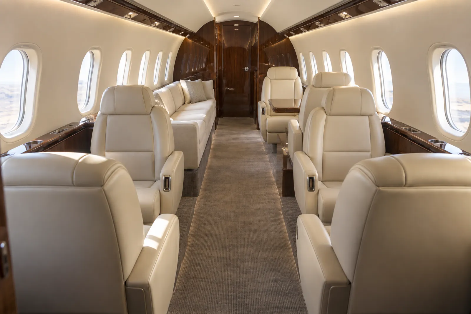 Warm natural light fills a Gulfstream G280 cabin viewed aft along its dropped aisle, with cream leather club seats, a three-place divan, upright rounded windows and polished wood veneer.