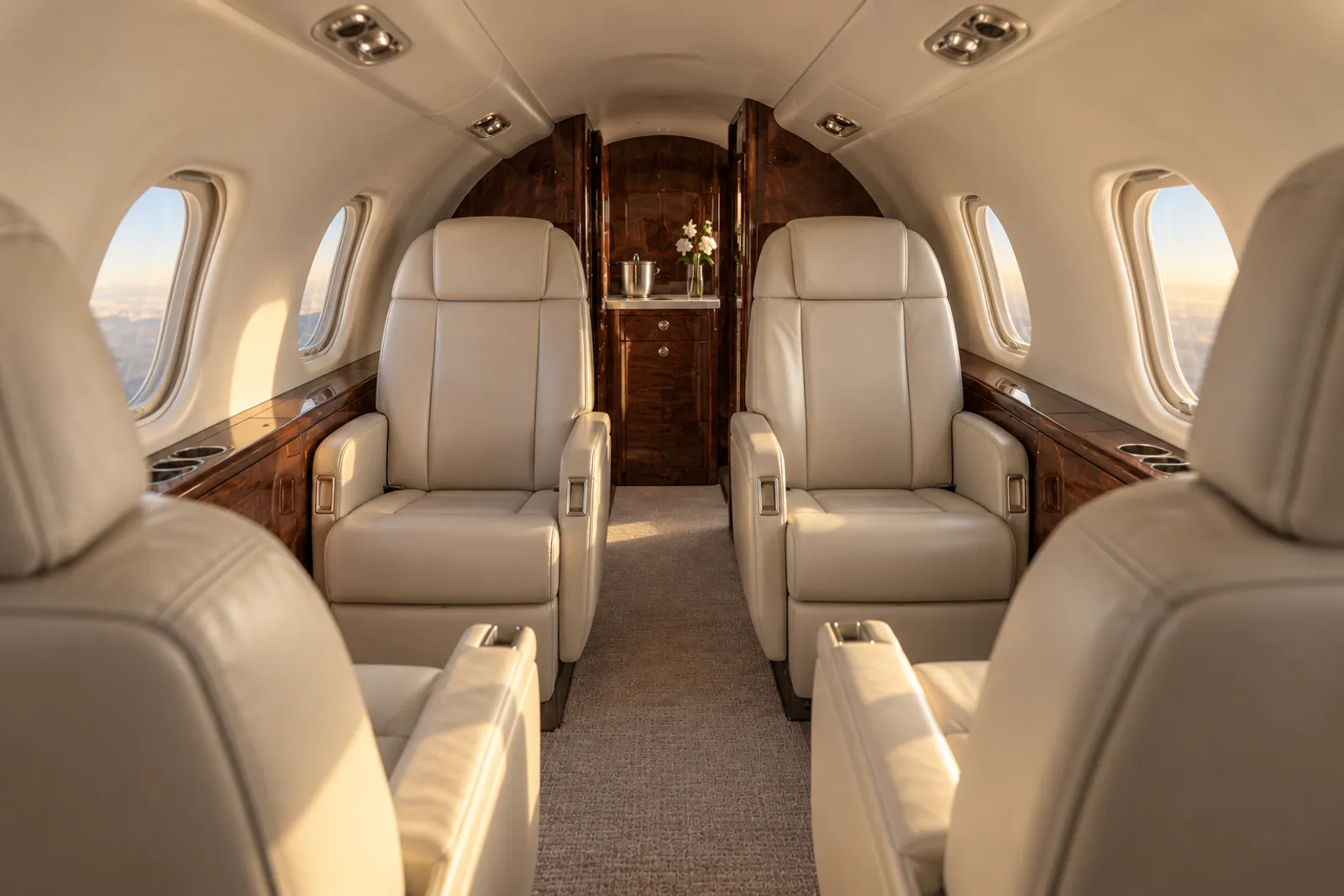 A warm, sunlit Learjet 45 cabin viewed aft along a narrow aisle, with a low curved ceiling, cream leather double-club seats, upright oval windows and polished wood trim.