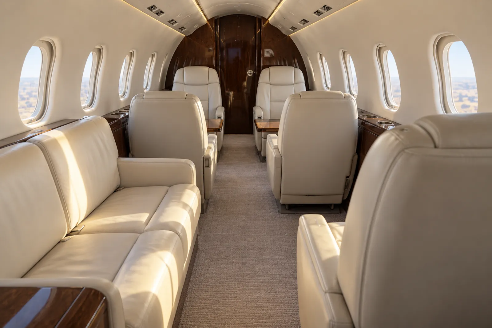An eye-level view looking aft through a Learjet 60 cabin shows cream leather chairs and a two-place divan, wood veneer, oval windows and a compact curved ceiling in warm natural light.