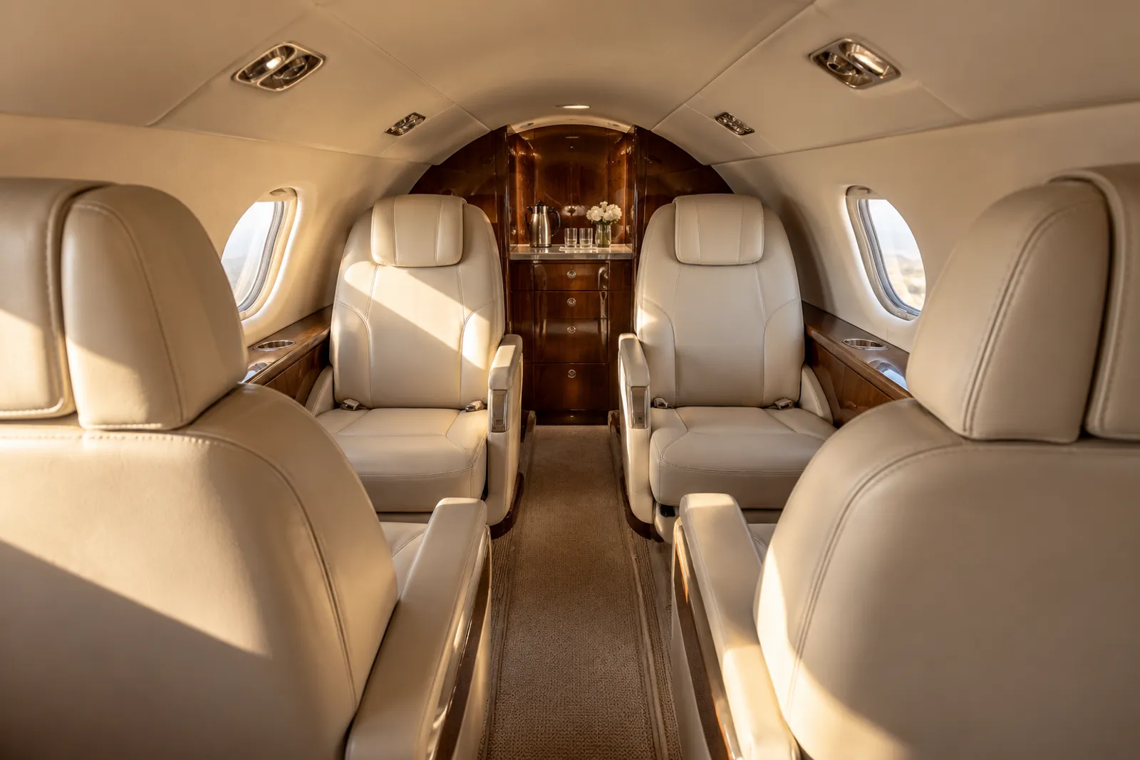 An eye-level view looking aft through a low Pilatus PC-24 cabin shows eight cream leather club seats, rounded rectangular windows and wood veneer in warm natural light.