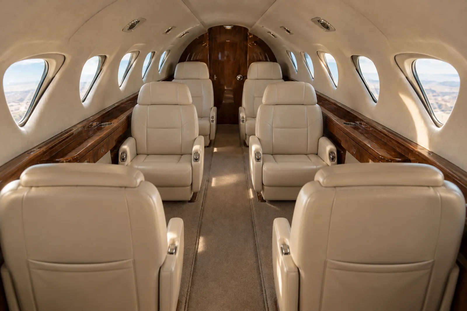 Warm sunlight illuminates six cream leather seats and wood veneer in the low, narrow Sabreliner 65 cabin, viewed aft along the aisle beneath a curved ceiling beside rounded triangular windows.